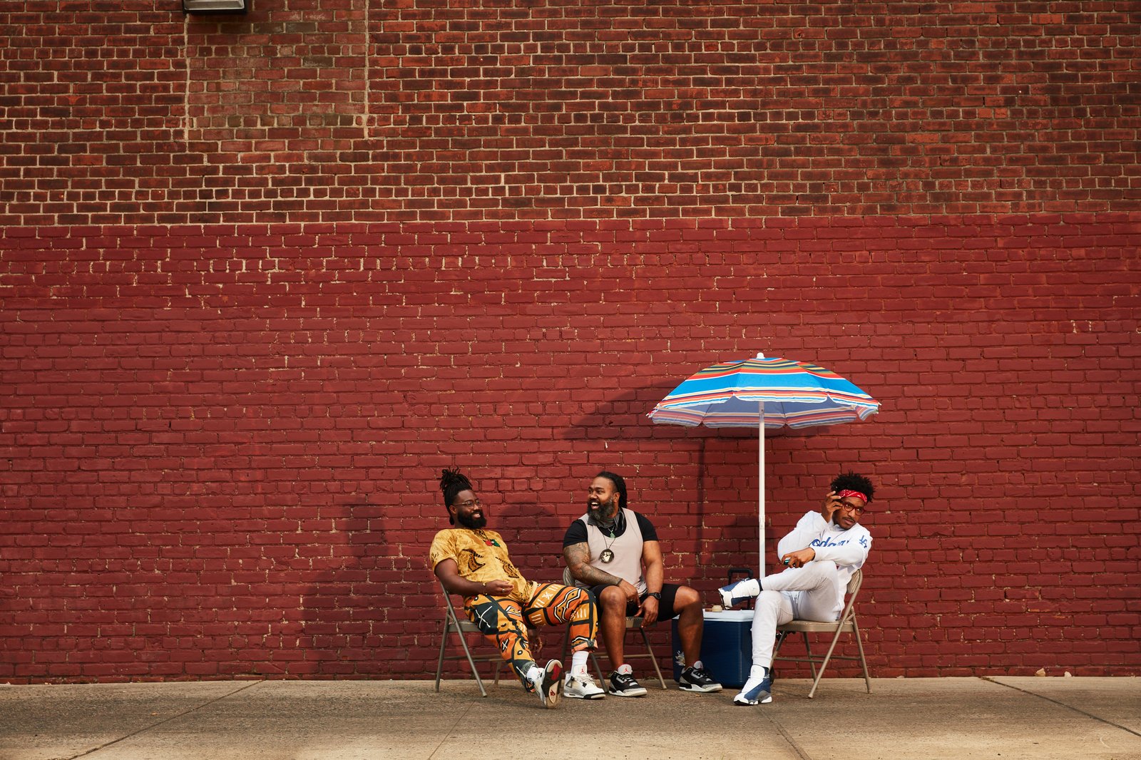 Three podcast co-hosts sitting outdoors under an umbrella against a red brick wall