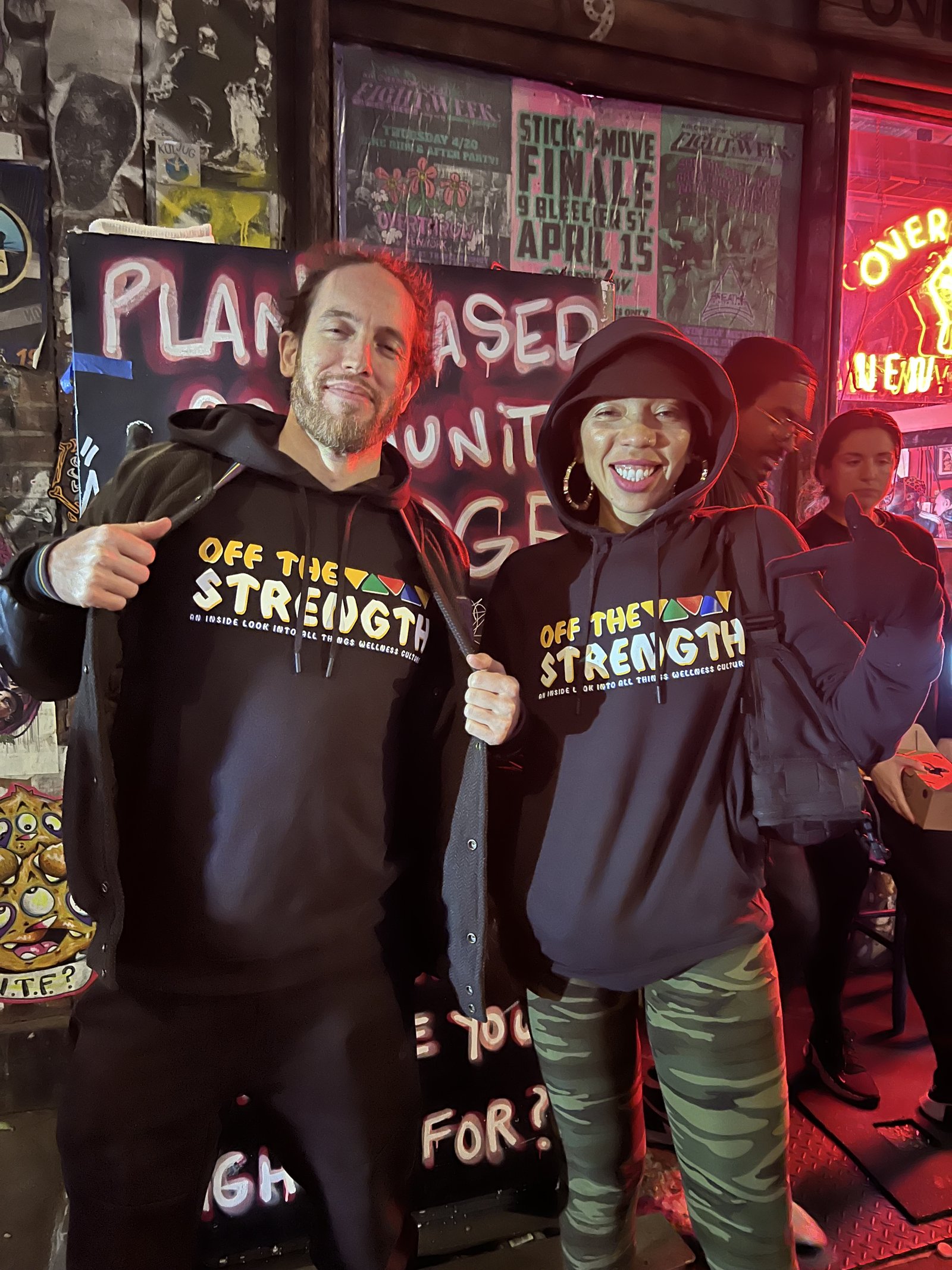 Two people wearing Off The Strength hoodies posing at a venue with show flyers
